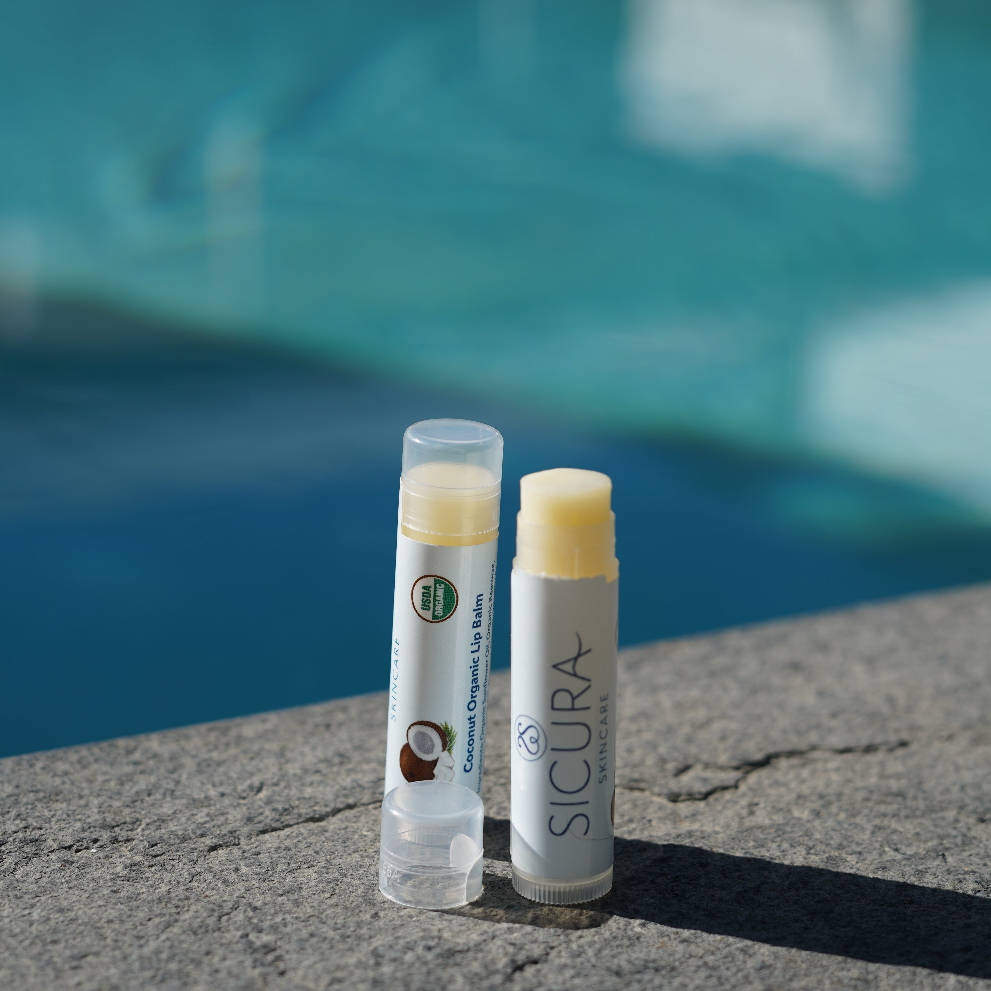 Two lip balm tubes labeled 'Sicura' on a poolside ledge with a blurred pool background.