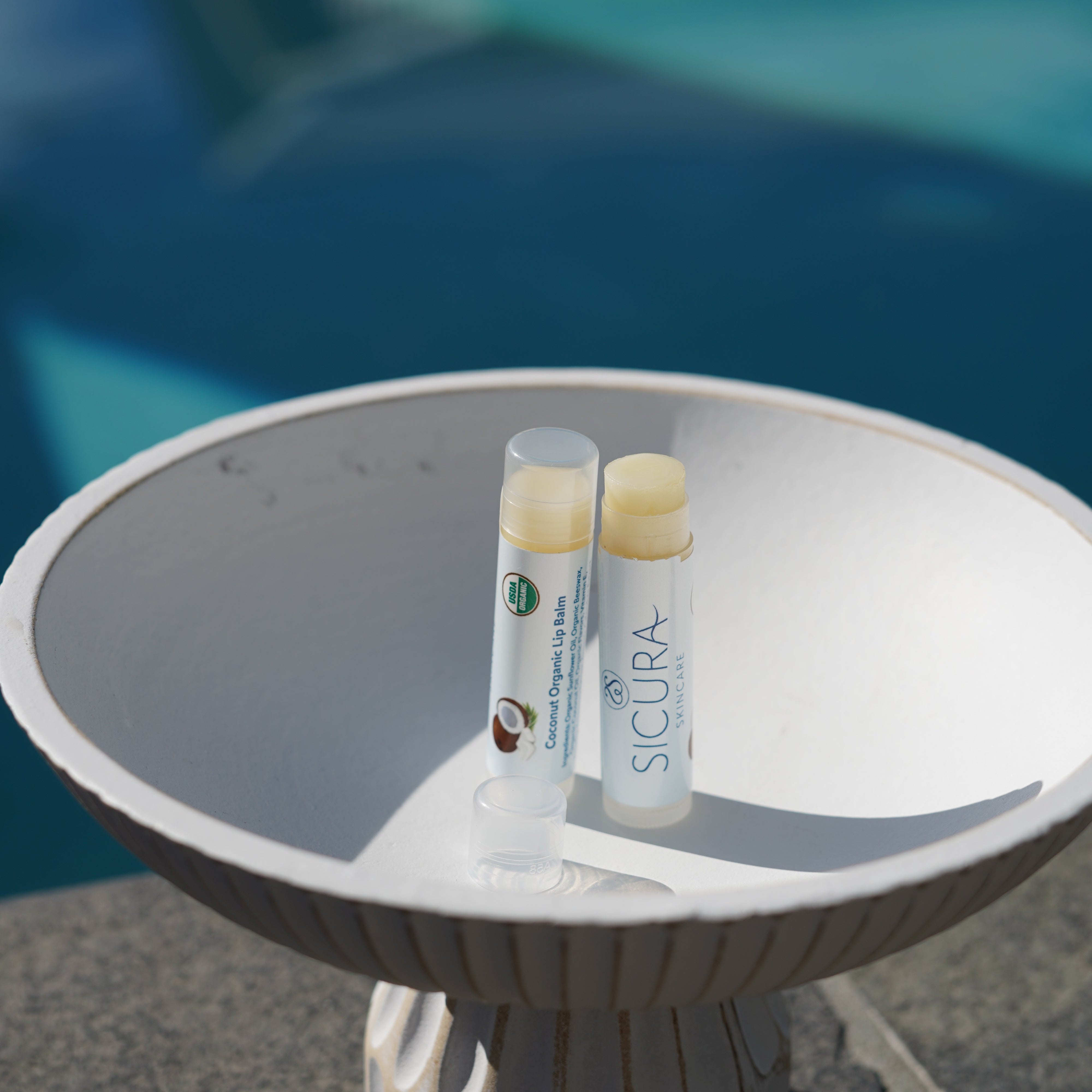 Two lip balms labeled 'SICURA' on a white surface with a blurred background