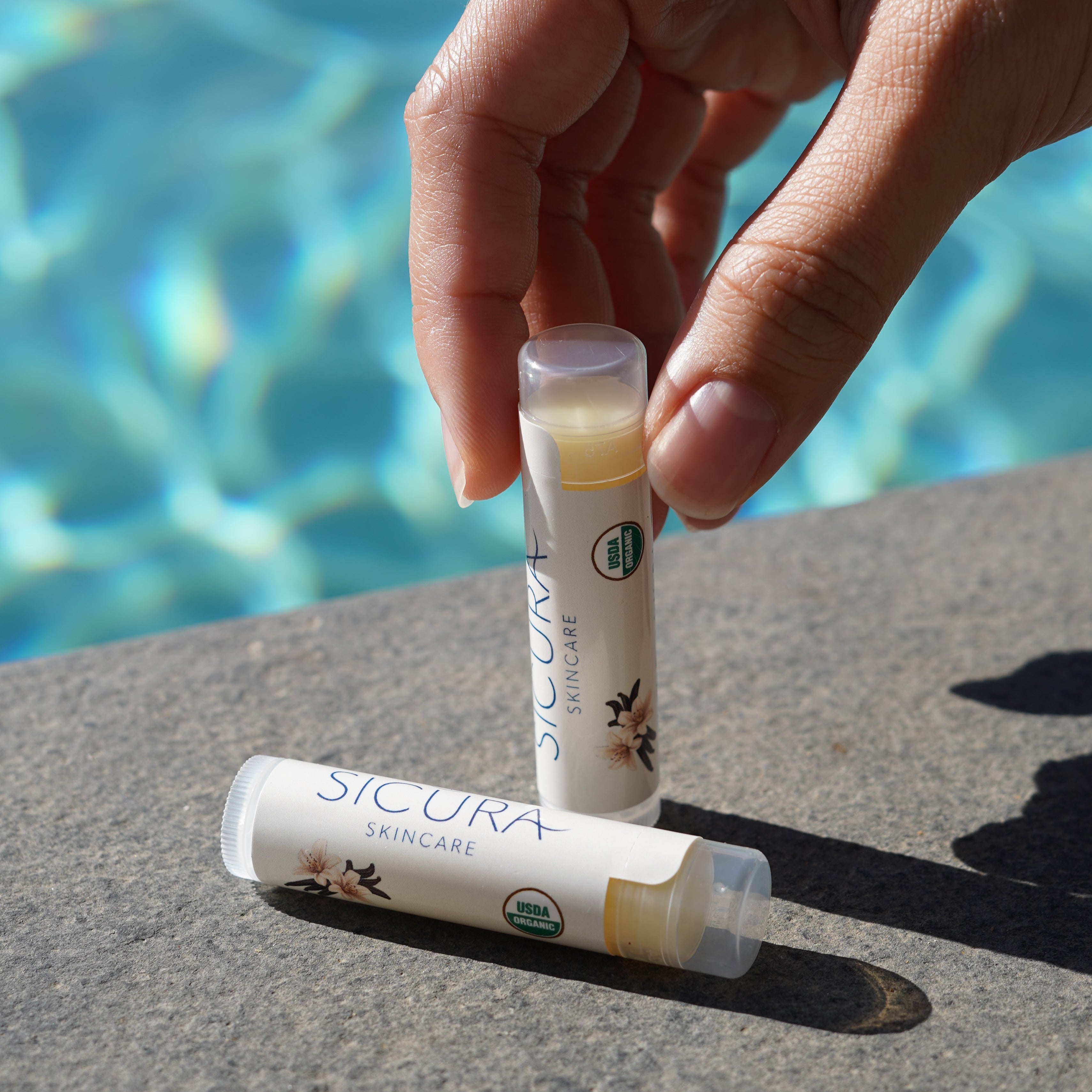 Hand holding two lip balm tubes with a pool in the background